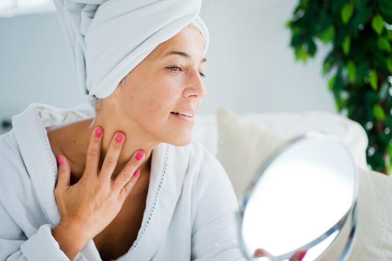 Woman looking herself into a mirror with towel on her head touching her neck with fingers while smiling